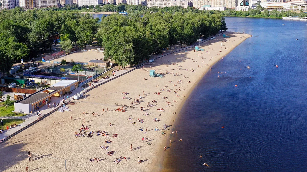 Пляж біля річки Дніпро у Києві, з людьми, що відпочивають на піску, і водою, що відбиває небо. Якість води на пляжах Києва